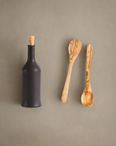 A handcrafted olive wood salad server set accompanied by a stoneware bottle, both displayed against a neutral background.