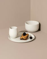 A minimalist white dinnerware set displayed against a neutral background, including a flat salad plate with a dessert, a dinner bowl, and a cup.