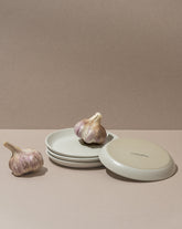 A white stoneware flat side plate with a matte finish, displayed alongside garlic bulbs. The plate is placed against a neutral background.