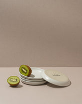 A white stoneware flat side plate with a matte finish, displayed with a halved kiwi on top, against a neutral background.