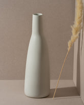 A white stoneware carafe with a narrow neck and a matte finish, displayed against a neutral background with a dry plant to the side.