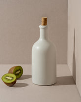 An olive oil bottle and a loaf of bread on a wooden board, with a small wooden bowl of olives beside it, all placed on a white surface.