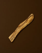 A wooden butter spreader with a curved handle, laying on a brown surface.