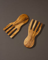A pair of flat olivewood serving forks placed on a dark surface.