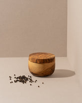 A round olive wood salt and spices cellar with a lid on a neutral background, surrounded by scattered spices.