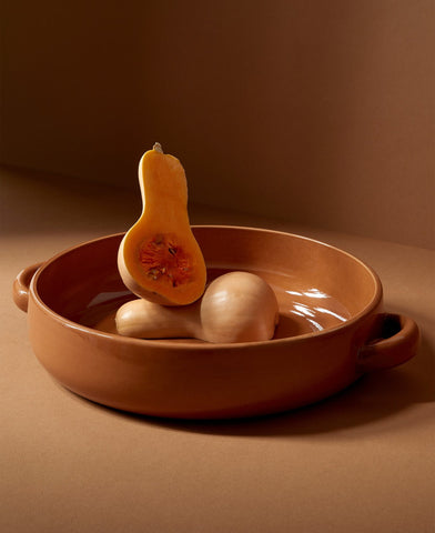 A beige stoneware serving platter with handles, containing what appears to be a roasted vegetable, placed on a brown surface.