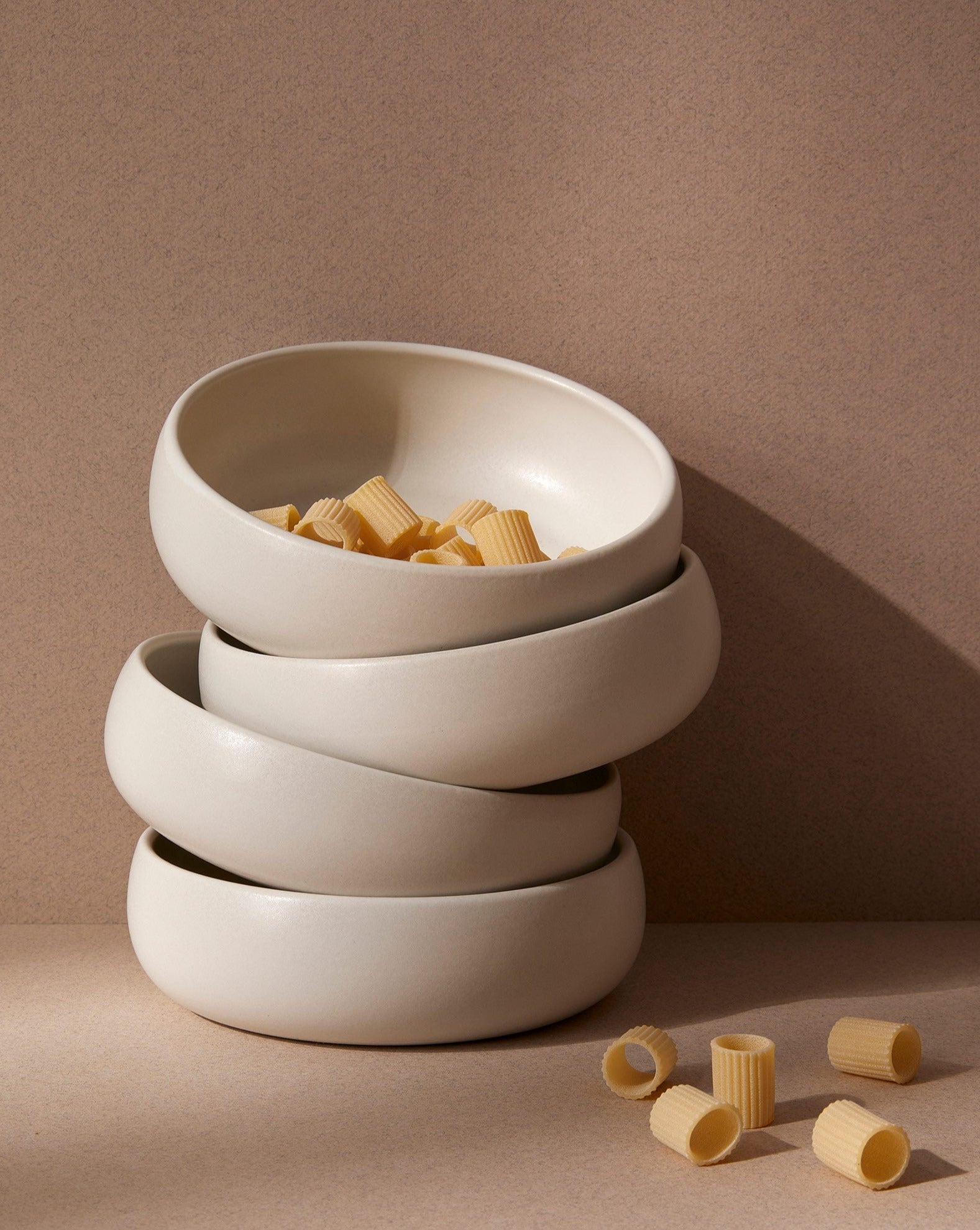 Stacked white stoneware tapas bowls with a beige rim, filled with what appears to be pasta. The bowls are set against a neutral background.