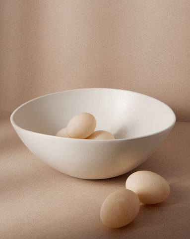 A matte white stoneware bowl with an egg inside, placed on an off-white background. Two additional eggs are positioned outside the bowl, creating a soft, minimal composition.