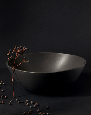 A matte black stoneware bowl placed on a dark surface, containing dried botanical elements with stems, creating a minimal and earthy aesthetic.