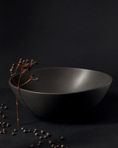 A black stoneware serving bowl with a matte finish on a neutral background, containing what appears to be a few eggs or round items, with the bowl's artisanal craftsmanship highlighted.