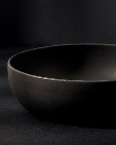 A matte black stoneware shallow serving bowl displayed against a dark background, showcasing its smooth, minimalist form and subtle texture.