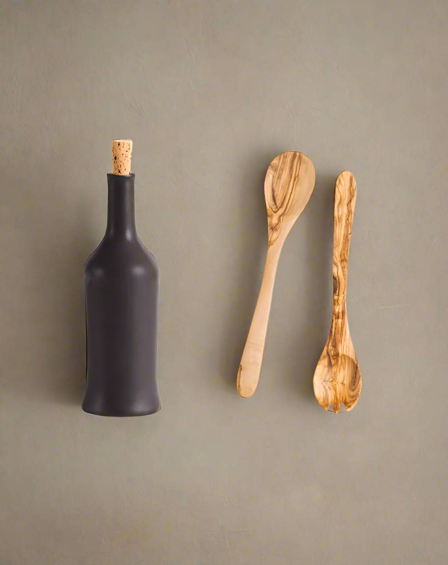A handcrafted olive wood salad server set accompanied by a stoneware bottle, both displayed against a neutral background.