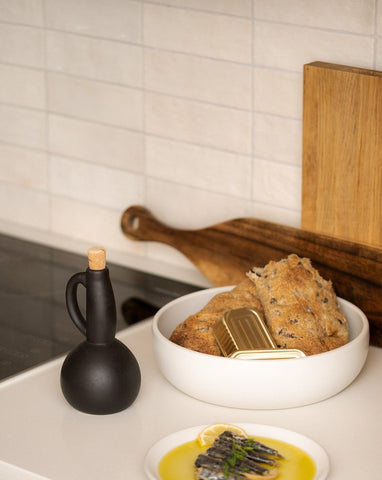 The same beige stoneware shallow serving bowl now filled with bread, placed beside a matching olive oil dispenser and a small flat plate holding a side dish, all arranged on a simple, neutral-toned surface.