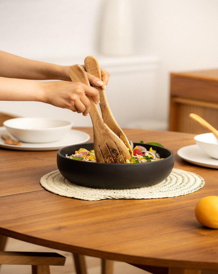 Handmade shallow stoneware bowl filled with salad, placed on a wooden dining table. A pair of natural olive wood serving spatula forks are shown mixing the salad, highlighting the organic textures and earthy, handcrafted design.