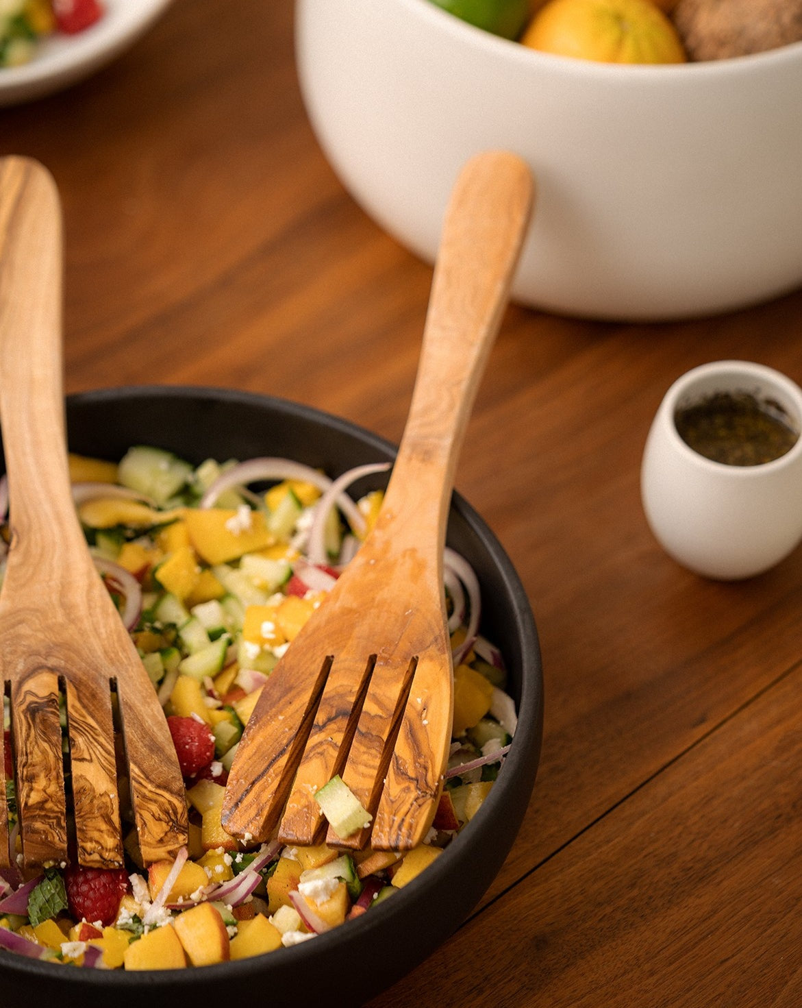 Natural olive wood serving spatula forks placed in a salad bowl, with a small cup and a large fruit bowl partially visible and blurred in the background.