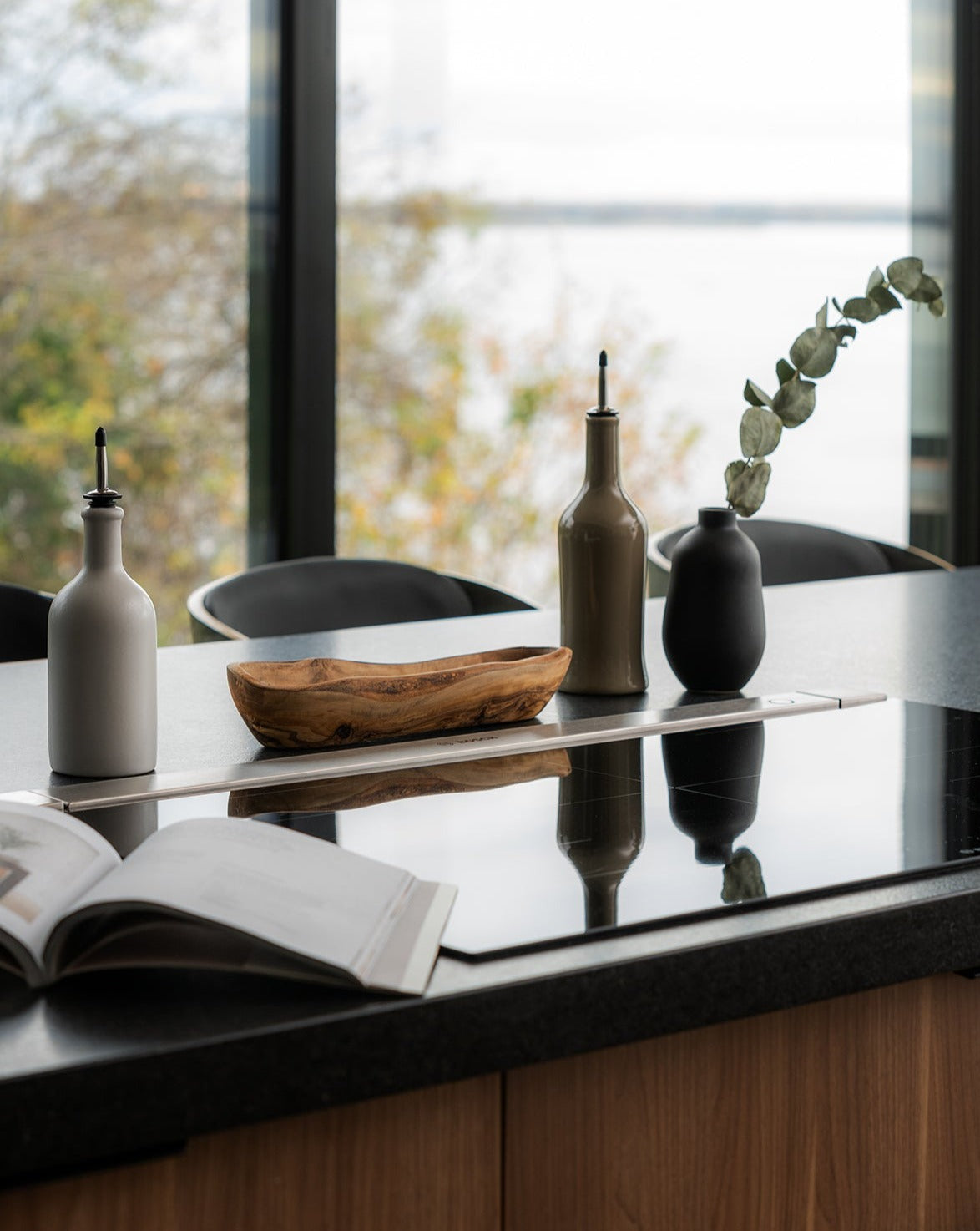 An empty oval-shaped rustic olive wood bowl displayed on a dining table. Three vases are arranged nearby, one holding a flower, and an open book rests at the edge of the table, creating a calm, styled setting.
