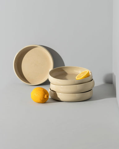 A beige stoneware pasta/salad plate with a contemporary round shape, displayed in a stacked arrangement with a white vegetable on the side.