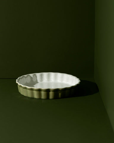 A white rounded stoneware pie dish with wavy side walls displayed on a neutral background, accompanied by a wooden cutting board with a pie on it, some garlic bulbs, and a small white bowl.