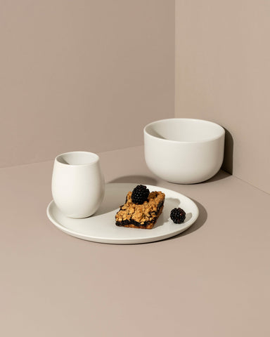 A minimalist white dinnerware set displayed against a neutral background, including a flat salad plate with a dessert, a dinner bowl, and a cup.