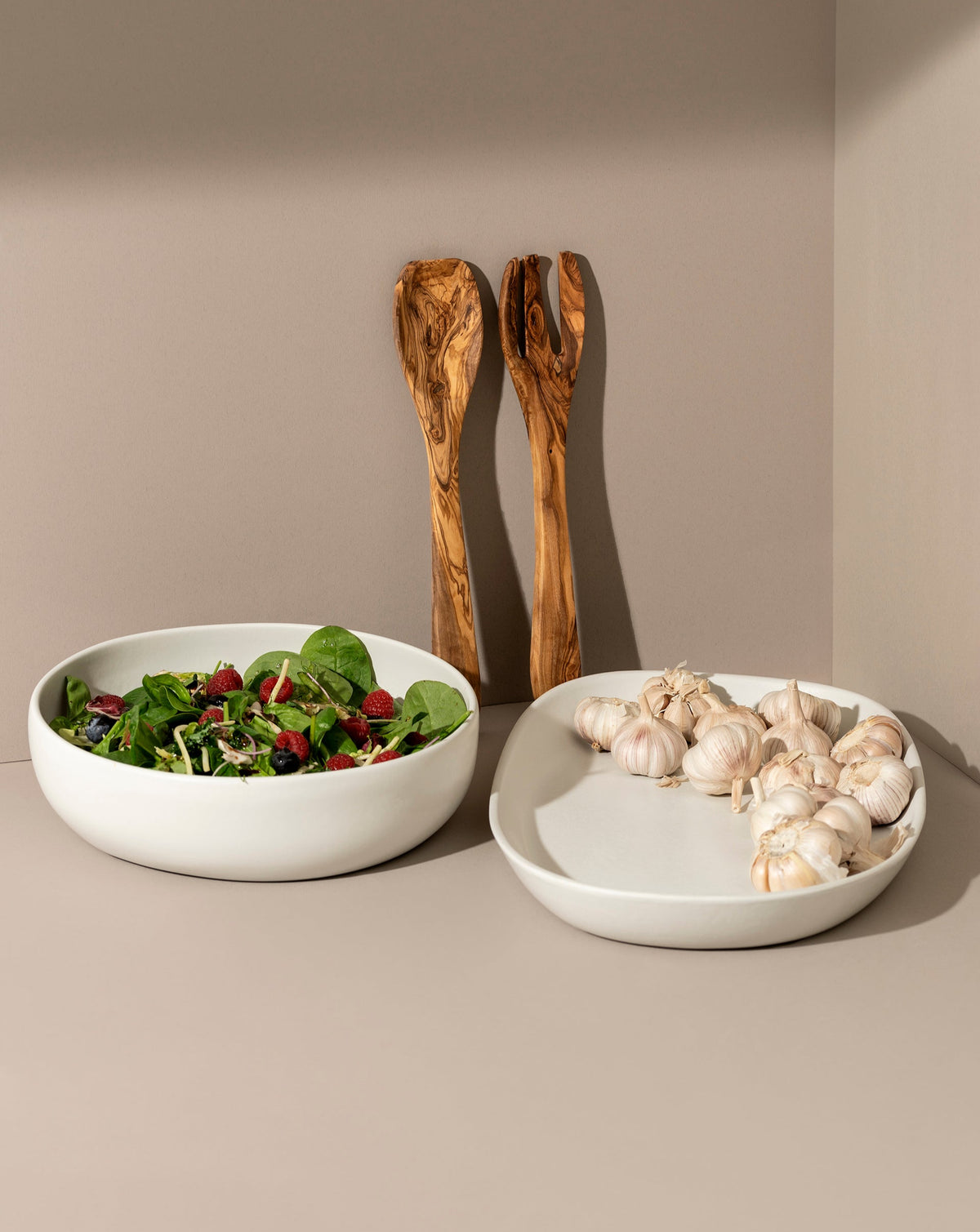 A white ceramic serving platter and bowl set with an olive wood serving set in the background, displayed with salad in the platter and garlic in the bowl.