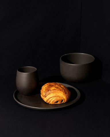 A minimalist white dinnerware set displayed against a neutral background, including a flat salad plate with a dessert, a dinner bowl, and a cup.