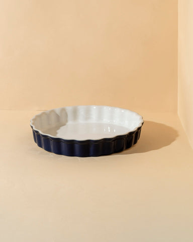 A white rounded stoneware pie dish with wavy side walls displayed on a neutral background, accompanied by a wooden cutting board with a pie on it, some garlic bulbs, and a small white bowl.
