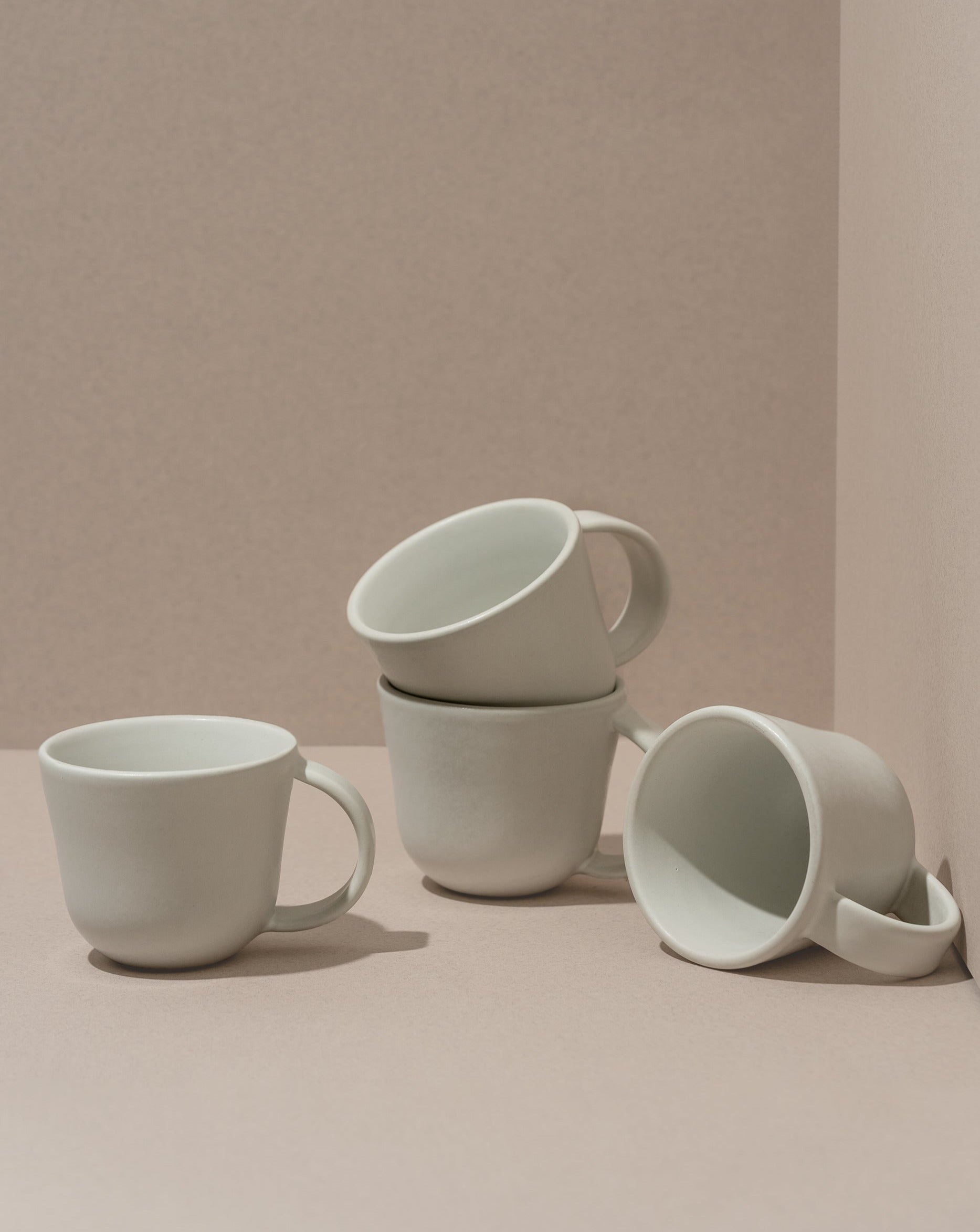 A set of four white stoneware coffee mugs, three stacked behind one in front, on a beige background.