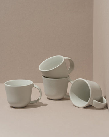A set of four white stoneware coffee mugs, three stacked behind one in front, on a beige background.