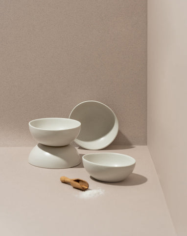A set of white stoneware condiment bowls with a matte finish, displayed against a neutral background. One bowl is upright, while the others are stacked behind it, and there is a small wooden spoon in front.