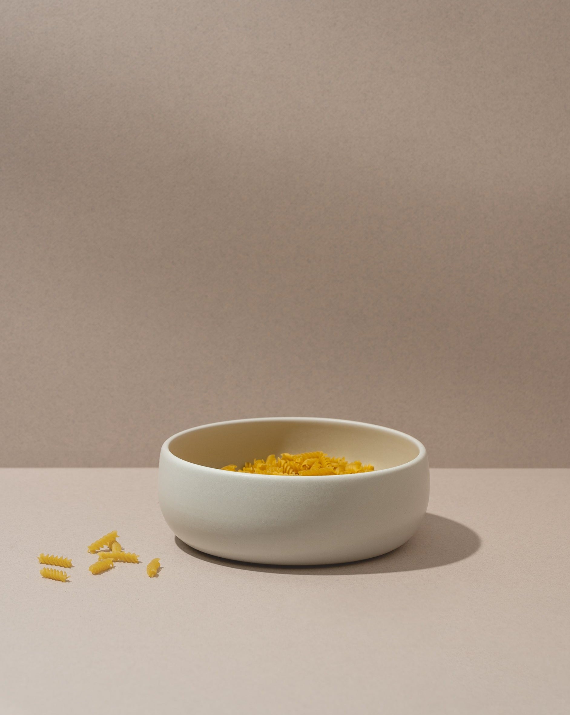 A white stoneware serving bowl with a simple design filled with yellow pasta. The bowl is placed on a neutral background.