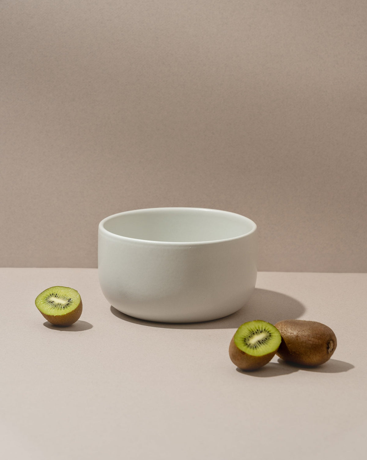 A white stoneware serving bowl on a neutral background with a kiwi cut in half beside it.