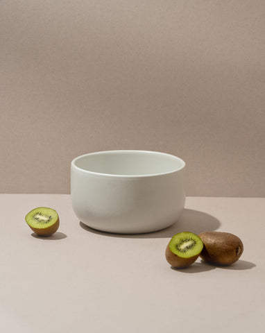 A white stoneware serving bowl on a neutral background with a kiwi cut in half beside it.