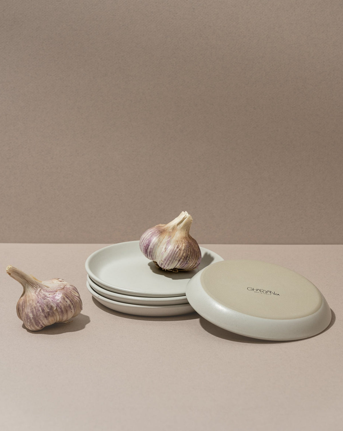 A white stoneware flat side plate with a matte finish, displayed alongside garlic bulbs. The plate is placed against a neutral background.
