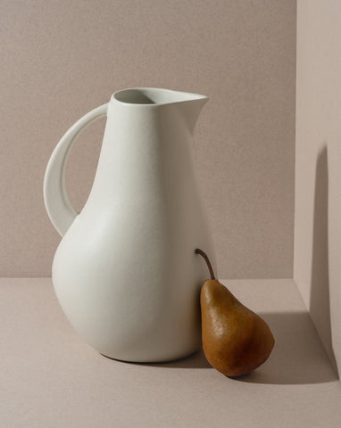 A white stoneware pitcher with a simple, elegant design, accompanied by a pear, presented against a neutral background.
