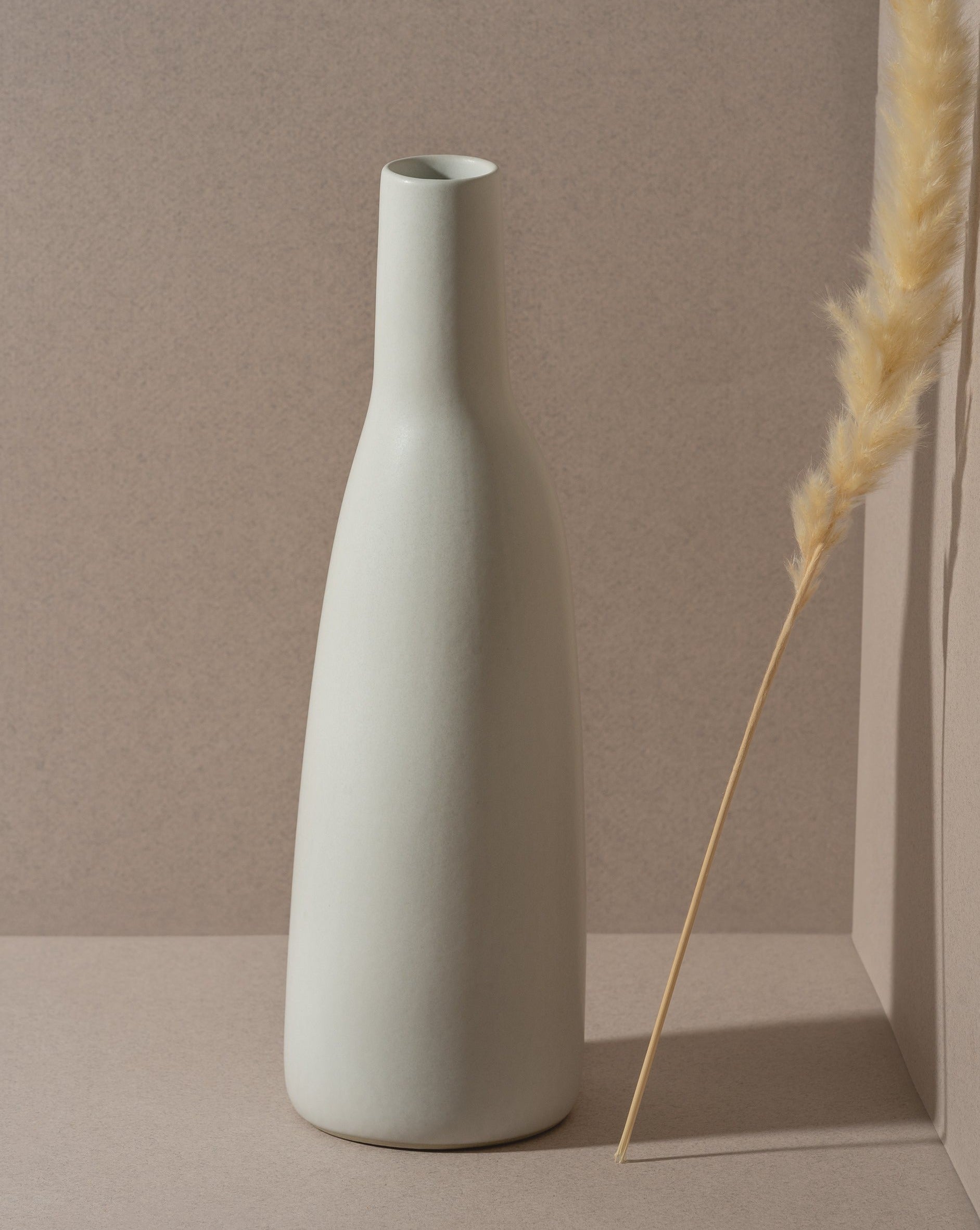 A white stoneware carafe with a narrow neck and a matte finish, displayed against a neutral background with a dry plant to the side.