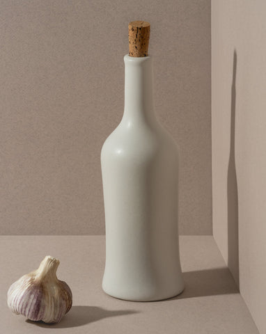 A white ceramic olive oil dispenser with a cork stopper, displayed next to a garlic bulb, on a neutral background.