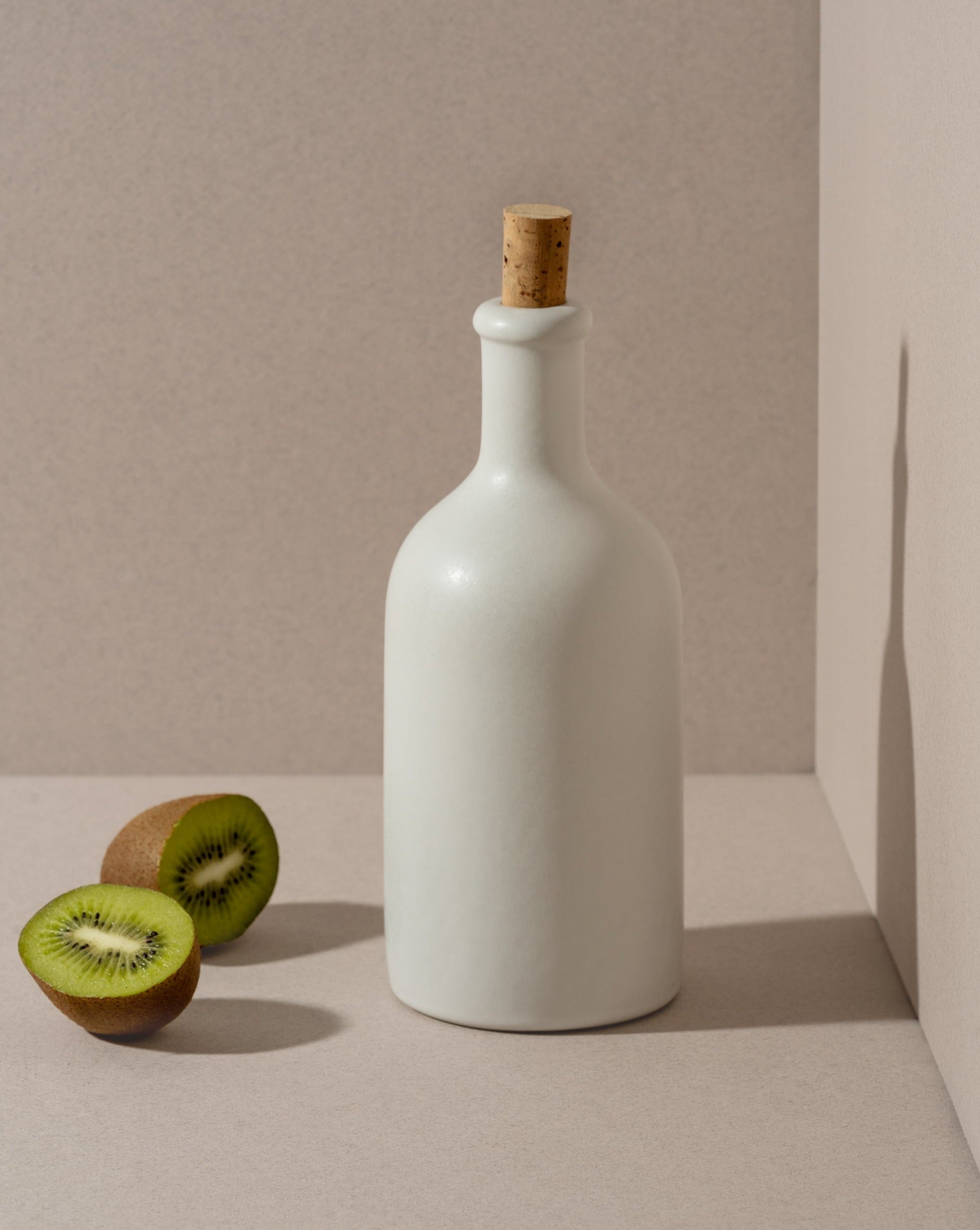 An olive oil bottle and a loaf of bread on a wooden board, with a small wooden bowl of olives beside it, all placed on a white surface.