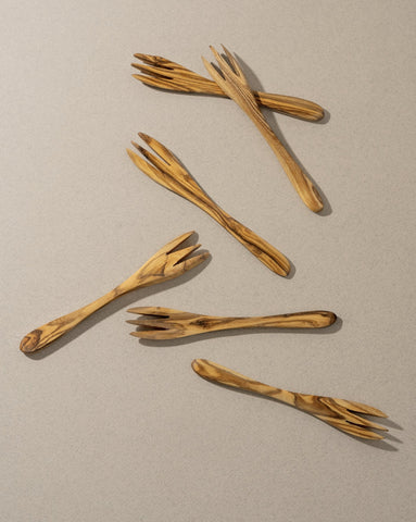 A set of natural olive wood tapas forks laid out on a grey surface.