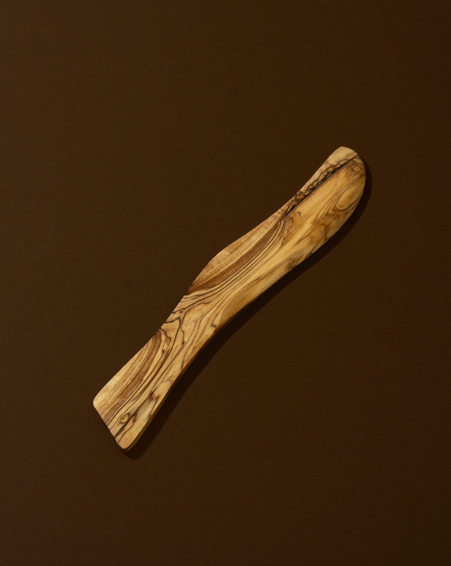 A wooden butter spreader with a curved handle, laying on a brown surface.