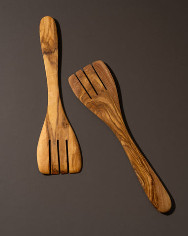 A pair of natural olive wood serving spatula forks laid on a dark surface.