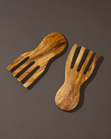 A pair of flat olivewood serving forks placed on a dark surface.