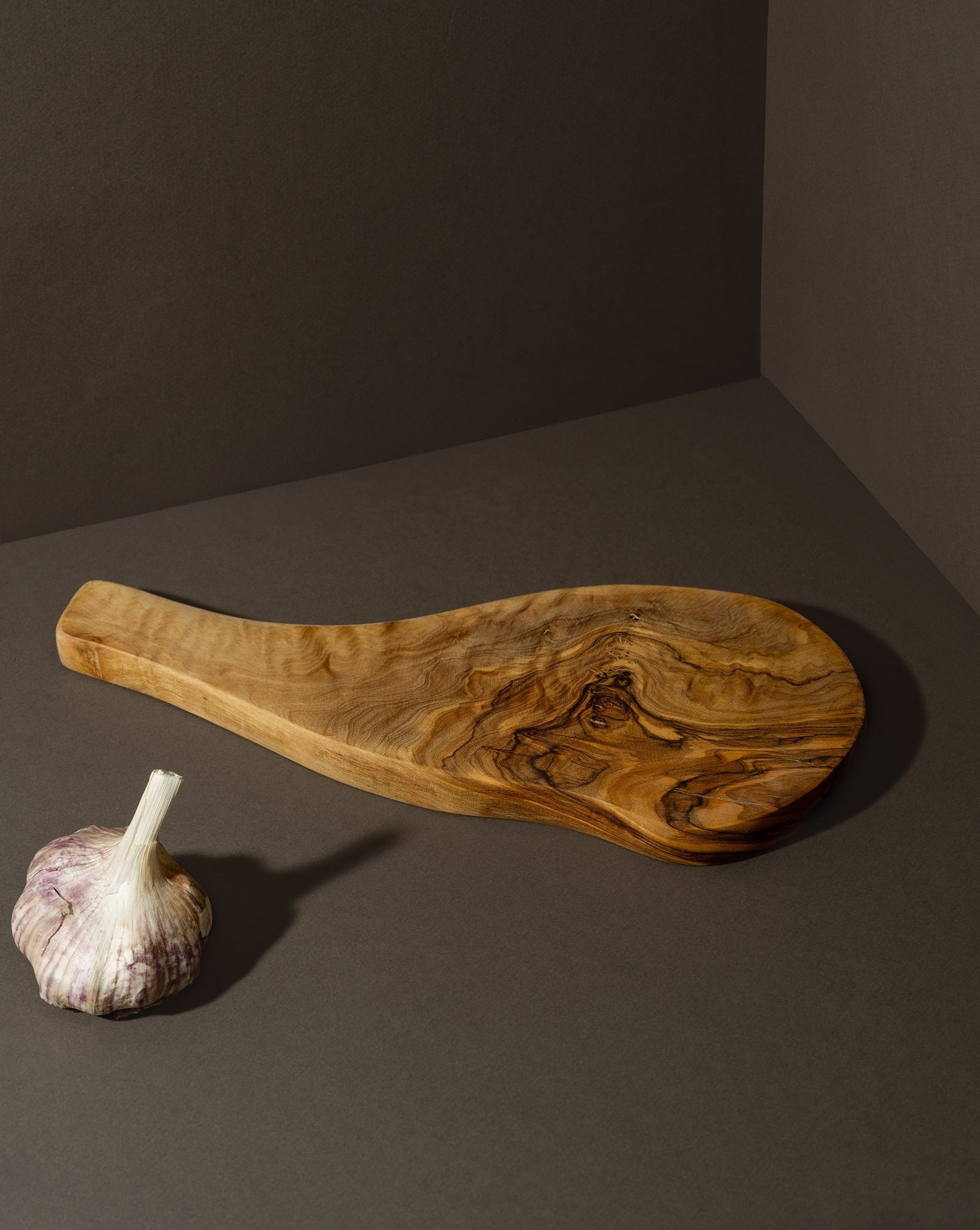 A natural olive wood cheese board with an organic shape and handle, displayed alongside a bulb of garlic.