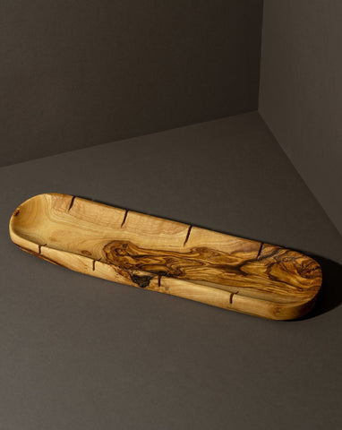 An olivewood sling bread board with visible natural cracks and imperfections, displayed on a dark background. Its unique shape and grain patterns highlight the rustic, handmade quality of the piece.