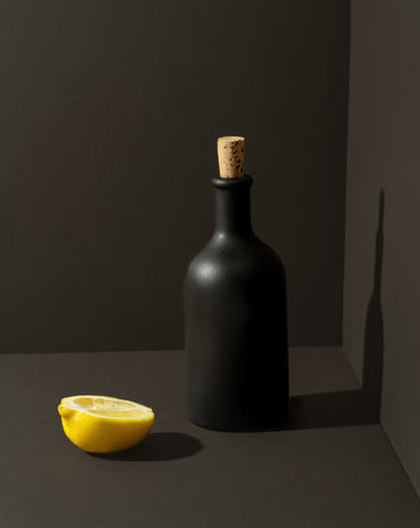 An olive oil bottle and a loaf of bread on a wooden board, with a small wooden bowl of olives beside it, all placed on a white surface.