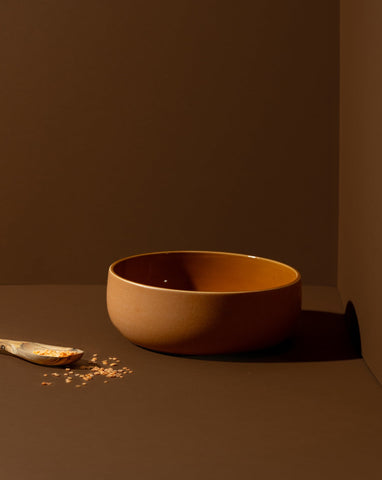 A terracotta-colored stoneware serving bowl placed on a brown surface. A wooden spoon rests beside it, with a few grains scattered around for a rustic touch.

