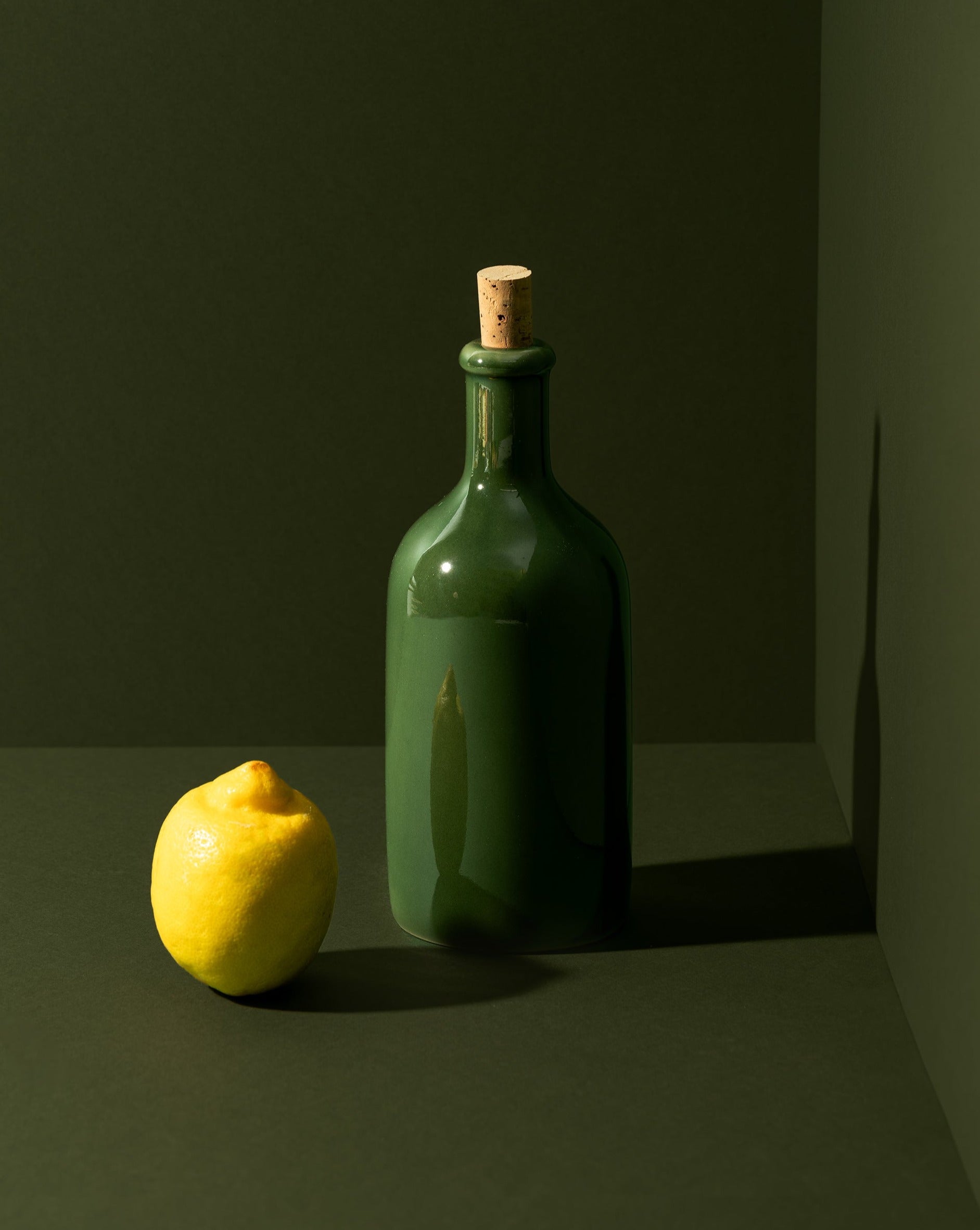 An olive oil bottle and a loaf of bread on a wooden board, with a small wooden bowl of olives beside it, all placed on a white surface.