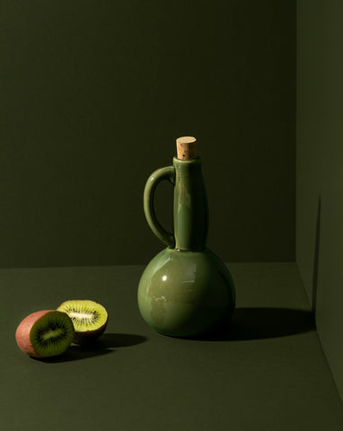 A white ceramic olive oil dispenser with a matte finish, a cork stopper, and a handle, displayed in a corner setting with nuts scattered in front.