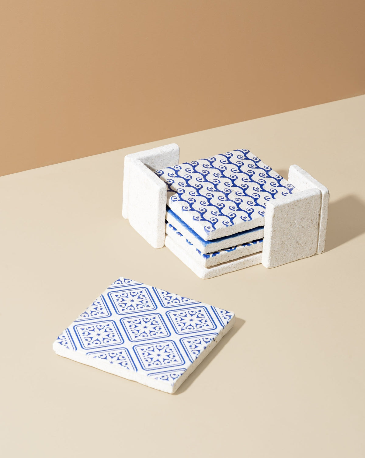 A set of four white marble coasters with blue patterned designs, displayed in a stacked formation with a beige holder.