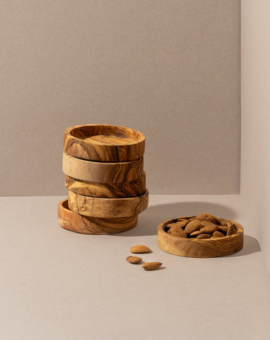 Five stacked olive wood bowls beside a separate bowl filled with walnuts, with a few scattered walnuts on the brown surface.