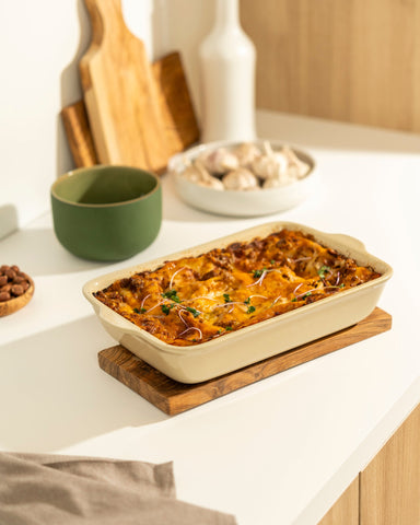 Rectangular beige stoneware baking dish filled with baked pasta, shown on a white surface with a Natural Olive Wood cheese board and a folded fouta towel placed beside it.